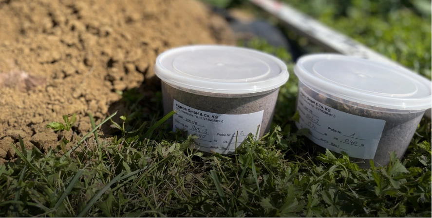 Mehrere durchsichtige Behälter mit Bodenproben stehen im Freien neben einer Erdgrube. Die Proben werden zur Untersuchung von Bodenqualität und Schadstoffen genutzt.