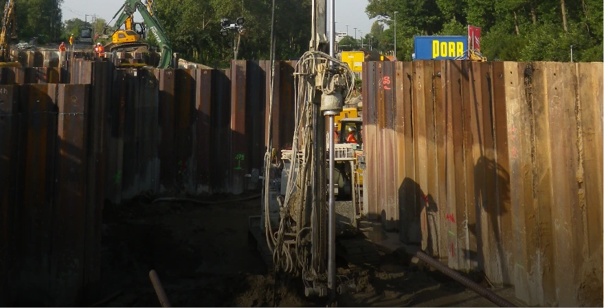 Auf einer Baustelle wird mit einem Bohrgerät und Spundwänden eine Baugrube gesichert, um trockene Arbeitsbereiche für Fundamentarbeiten zu schaffen.