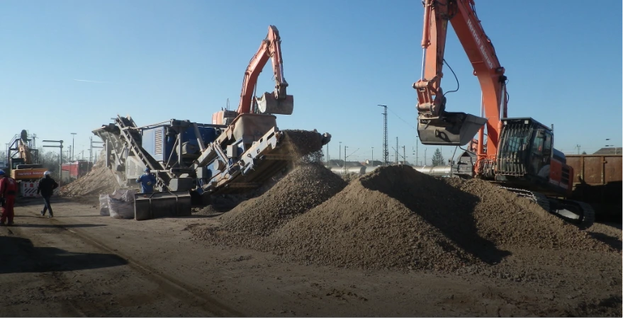 Zwei Bagger und eine Brechanlage zerkleinern Bauschutt auf einer Baustelle. Arbeiter trennen und überwachen das Material für eine umweltgerechte Entsorgung.