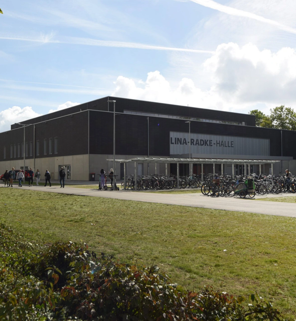 Moderne Sporthalle mit dunkler Fassade, großen Fenstern und Fahrradständern im Vordergrund.