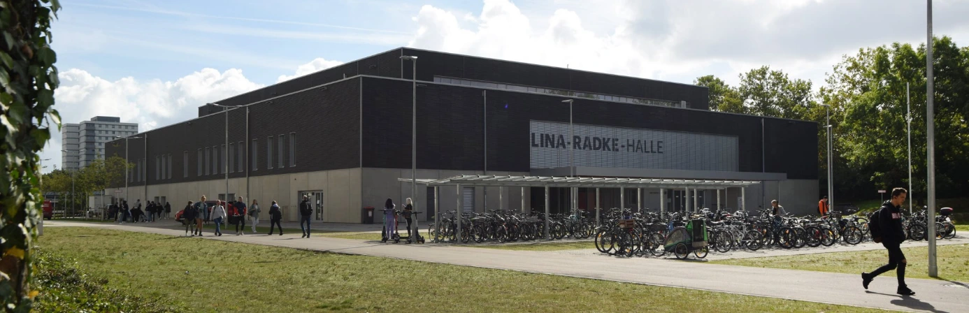 Moderne Sporthalle mit dunkler Fassade, großen Fenstern und Fahrradständern im Vordergrund.