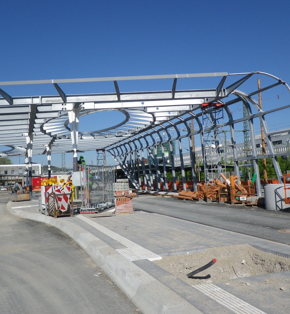 Baustelle mit moderner Stahlkonstruktion für eine neue Haltestellenüberdachung. Materialien und Baugeräte sind vor Ort sichtbar.